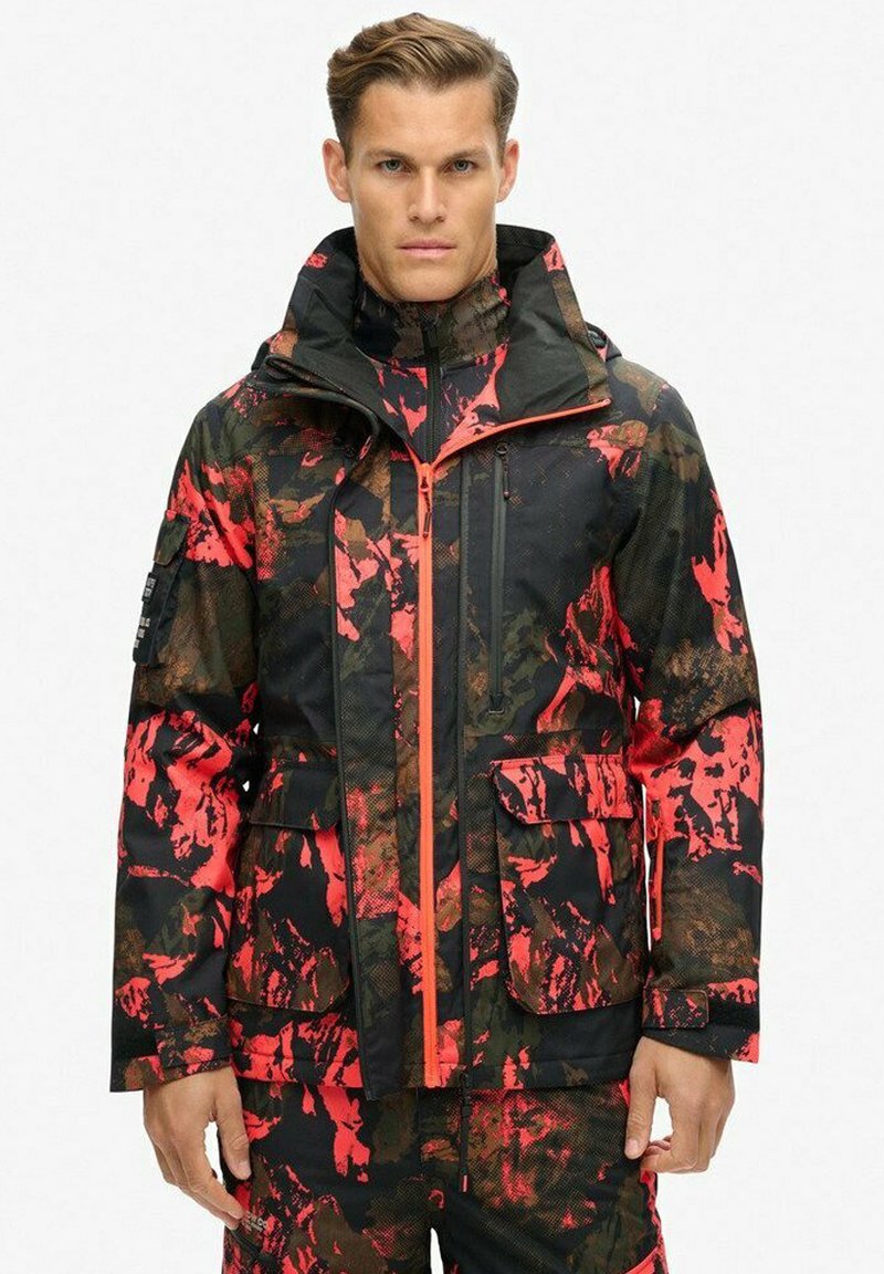 Chaqueta impermeable con una base negra y patrones abstractos en rojo y marrón, con cierre delantero, capucha ajustable y múltiples bolsillos.