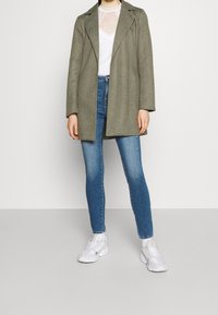 Olivově zelený kabát z vlněné směsi s klopami, nošený přes bílou síťovanou halenku s puntíky, spárovaný s modrými skinny džíny a bílými teniskami.