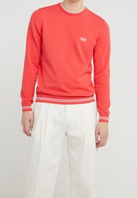 Roter Rundhalsausschnitt-Pullover mit weißem Logo, gerippten Bündchen und Saum, die zwei weiße Streifen aufweisen, kombiniert mit weißen plissierten Hosen.