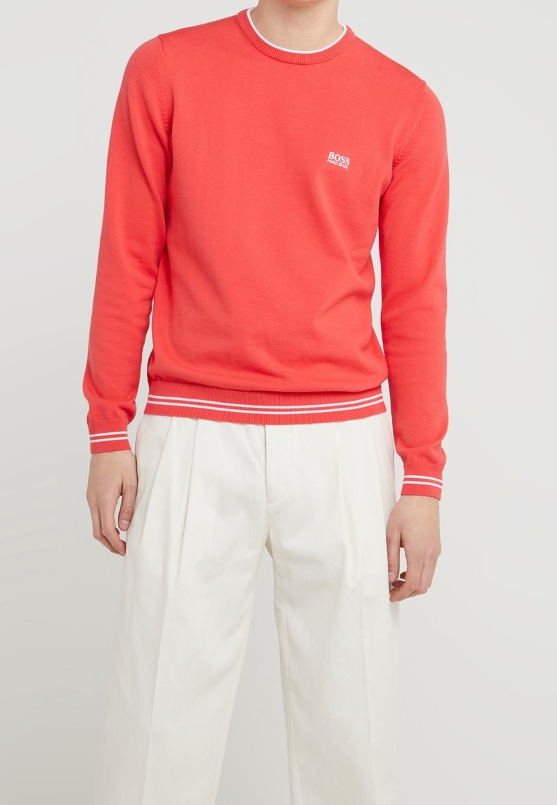 Roter Rundhalsausschnitt-Pullover mit weißem Logo, gerippten Bündchen und Saum, die zwei weiße Streifen aufweisen, kombiniert mit weißen plissierten Hosen.
