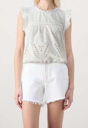 White sleeveless blouse with eyelet embroidery and ruffle shoulders paired with white frayed hem denim shorts on a standing person.