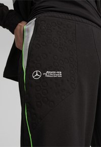 Zwarte sportieve broek met een gestructureerd patroon, groene biezen, ritszak en wit AMG Petronas Formule 1 Team-logo op de linker dij.