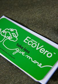 Grön EcoVero-tag på en texturerad mörk tygbakgrund. Texten inkluderar "ta mindre" och "ge mer" med en trägrafik.