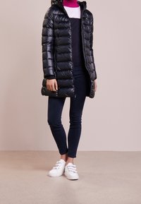 Abrigo negro tipo puffer con capucha, con secciones acolchadas, combinado con jeans pitillo oscuros y zapatillas blancas. Textura lisa, diseño de longitud media.