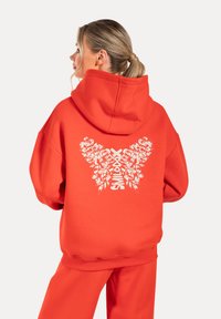Sudadera roja con un gran diseño de mariposa blanca que presenta elementos florales en la parte posterior. Tiene una capucha con cordón y puños de canalé.