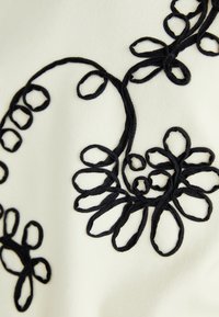 Svart brodert blomstermønster på et lys kremfarget stoff, med sløyfer og kurver, med varierende størrelser og former på kronbladene.