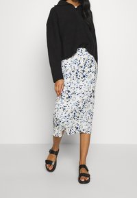 Haut en tricot noir court avec fermeture éclair, associé à une jupe midi florale blanche avec des accents bleus et jaunes, et des sandales noires.