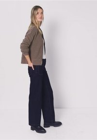 Cardigan de rayas marrón y crema con textura acanalada, combinado con vaqueros oscuros de corte holgado y botines negros. Posición de pie.