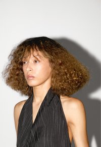 Zwarte halterjurk met verticale streepjes; model heeft volumineus, krullend haar; zachte belichting creëert een subtiele schaduweffect.