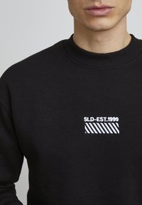 Zwarte sweatshirt met een ronde halslijn, voorzien van witte geborduurde tekst "SLD-EST.1999" en een gestreept patroon op het linkerbovenbeen.