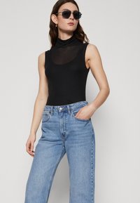 Black ribbed bodysuit with a high neckline, paired with light blue wide-leg jeans. Model wears dark sunglasses and has shoulder-length hair.