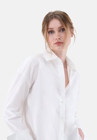 Weißes Button-down-Hemd aus glattem Stoff, mit klassischem Kragen, langen Ärmeln und lockerem Schnitt mit seitlichen Taschen.