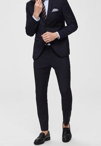 Mies, kuris vilki tamsiai mėlyną kostiumą su dryžuota kaklaraiščiu, baltu marškiniu, raštuota kišenine ir juodais odiniais loafer'iais ant vientiso fono.