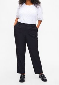 Femme portant un blouson court à manches courtes texturé blanc, un pantalon droit noir et des mocassins noirs brillants, se tenant avec les mains dans les poches.