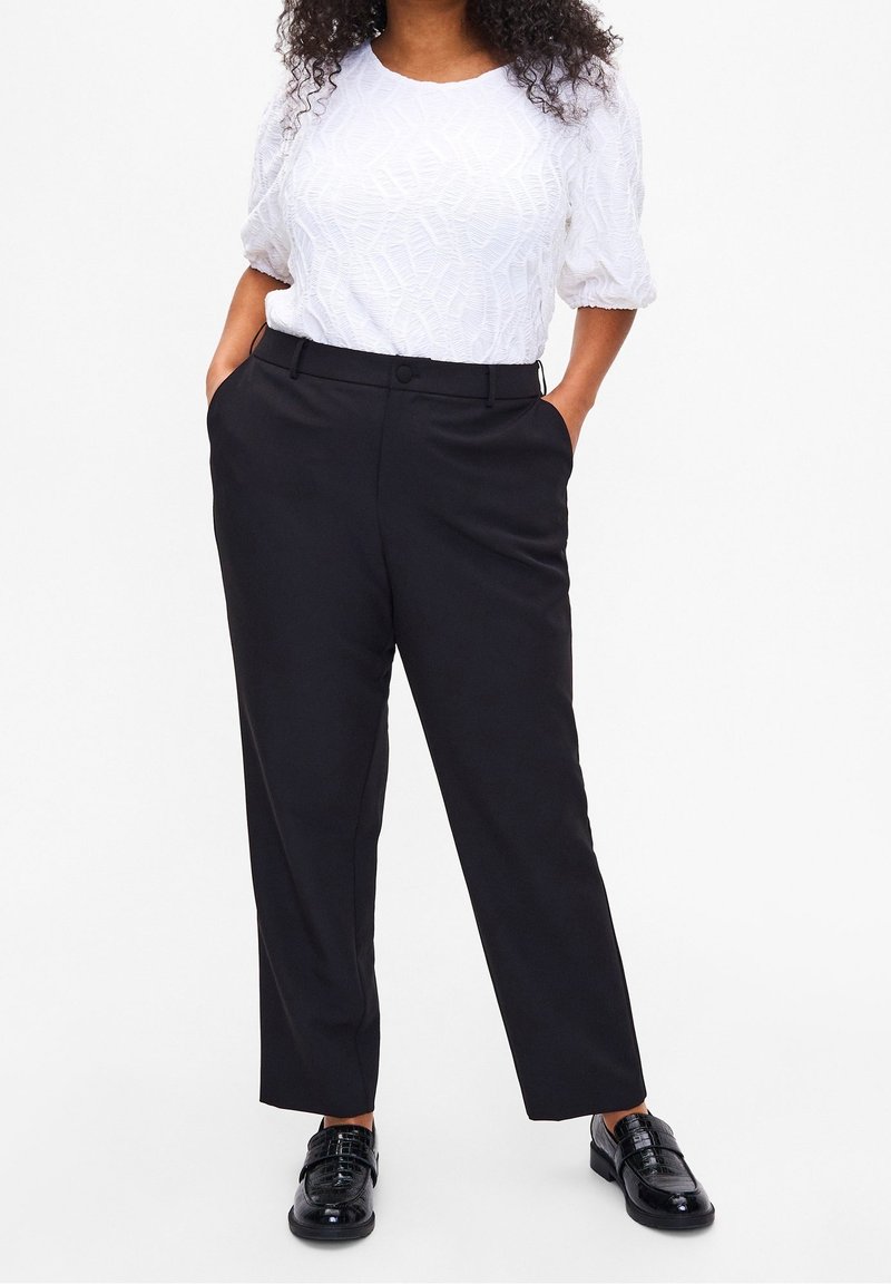 Femme portant un blouson court à manches courtes texturé blanc, un pantalon droit noir et des mocassins noirs brillants, se tenant avec les mains dans les poches.