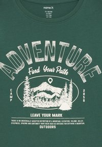 T-shirt de algodão verde escuro com texto gráfico branco e design de montanha. O texto inclui "AVENTURA", "Encontre o Seu Caminho" e imagens de acampamento.