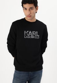 Fekete pulóver hosszú ujjakkal és kerek nyakkivágással. Elülső részén egy vastag, csíkos betűtípussal a "KARL LAGERFELD" felirat látható.