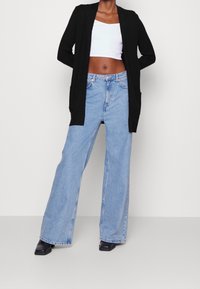 Cardigan noir ouvert sur un crop top blanc, associé à un jean bleu clair à jambes larges. La tenue comprend deux poches avant et des chaussures noires à bouts carrés.