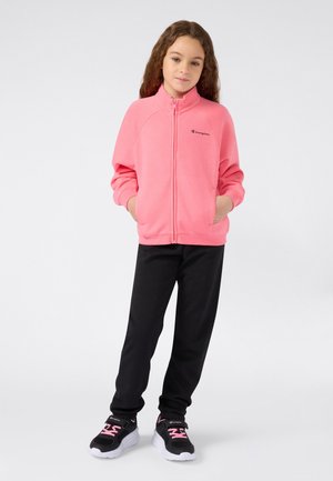 Meisje met lang krullend haar draagt een roze zip-up jacket, zwarte joggingbroek en zwarte sneakers met roze veters op een witte achtergrond.