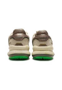 Un paio di sneakers beige e marroni con suole bianche spesse e suole esterne testurizzate di colore verde brillante, mostrato da dietro su sfondo bianco.