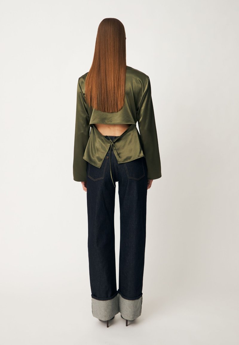 Personne aux longs cheveux raides portant un top en satin vert olive noué dans le dos et un jean large foncé avec de grands revers roulés et des talons hauts.