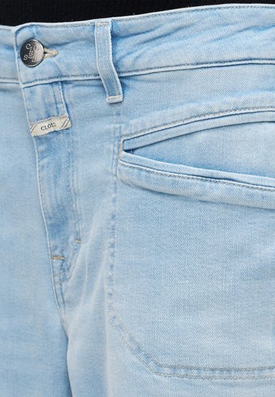 Ανοιχτό μπλε τζιν παντελόνι σε κοντινό πλάνο που δείχνει την μπροστινή τσέπη, την θηλιά για ζώνη και το μεταλλικό κουμπί με το όνομα της μάρκας «Closed».