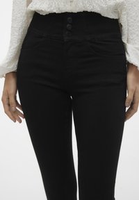 Jeans negros de cintura alta con un diseño ajustado, cierre de tres botones y bolsillos frontales, fabricados con una tela elástica de textura suave.