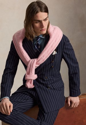 Mann mit schulterlangem Haar in einem marineblauen Nadelstreifenanzug, blauem Hemd und gestreifter Krawatte, mit einem rosa Pullover über den Schultern.