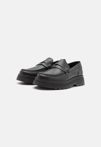 Mocassins en cuir noir avec détails cousus, un design élégant, et une semelle en caoutchouc épaisse et texturée pour une meilleure adhérence et durabilité.