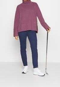 Übergroßer lila Rollkragenpullover mit langen Ärmeln, kombiniert mit schmales, navy Hosen und weißen Sneakern, während ein Golfschläger gehalten wird.