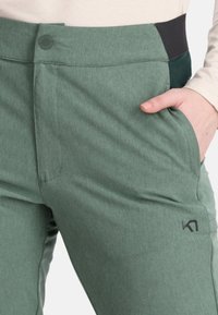 Pantalon sportif vert en tissu texturé, avec une ceinture élastique noire, des poches latérales et un petit logo sur la cuisse.