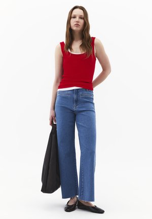 Junge Frau steht, trägt ein rotes Tanktop, blaue weit geschnittene Jeans, schwarze Ballerinas und hält eine große schwarze Tragetasche vor weißem Hintergrund.