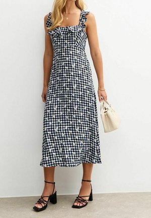 Robe vichy bleu foncé et blanche avec des bretelles à volants, des détails floraux, de longueur midi, assortie à des talons noirs à lanières et un sac à main crème.