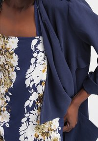 Marinblus som är lager på lager över en blommönstrad klänning med vita och gyllene accenter. Blusen har en avslappnad passform och en mjuk textur.