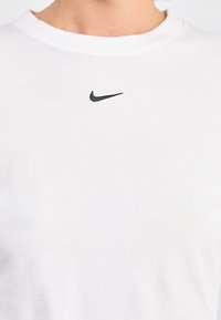 Hvit bomulls t-skjorte med rund hals, med en svart brodert Nike swoosh-logo i midten. Glatt tekstur, enkelt design.