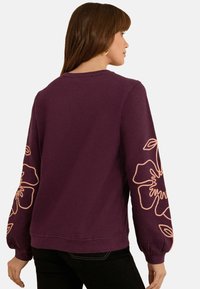 Sweatshirt violet à manches ballon, avec un motif floral en pêche sur les deux manches. Texture lisse, col rond.