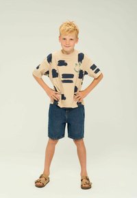 Camiseta de manga corta beige con un patrón de pinceladas azul marino, complementada con pantalones cortos de mezclilla y sandalias marrones. Diseño sencillo con un corte holgado.
