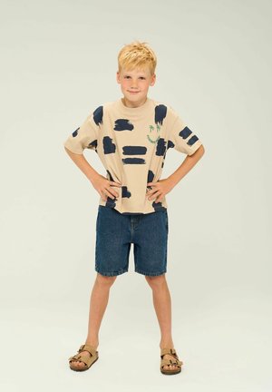 Beige kortärmad t-shirt med marinblå borststrykmönster, kompletterad med jeansshorts och bruna sandaler. Enkel design med avslappnad passform.