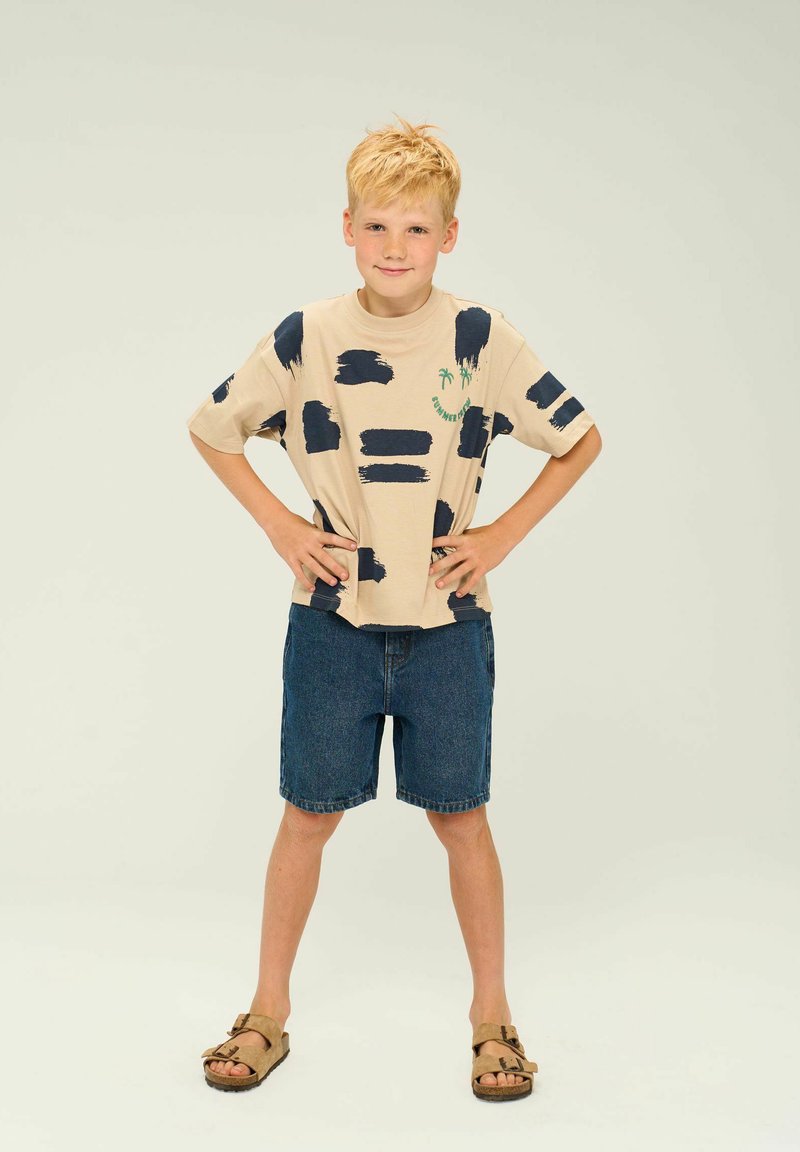 Camiseta de manga corta beige con un patrón de pinceladas azul marino, complementada con pantalones cortos de mezclilla y sandalias marrones. Diseño sencillo con un corte holgado.