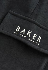Čierna látková etiketa s nápisom "BAKER BY TED BAKER" v bielych vystúpených písmenách, s hladkou textúrou a obdĺžnikovým tvarom.