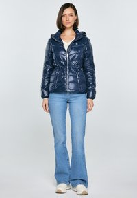 Kobieta ubrana w granatową pikowaną kurtkę z troczkiem w talii, jasnoniebieskie dzwoniaste dżinsy oraz białe tenisówki, stojąca na jednolitym tle.