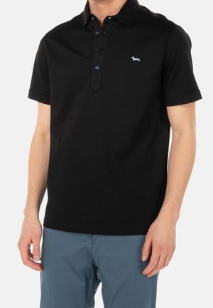 Homme portant un polo noir à manches courtes avec un petit logo de chien sur le côté gauche de la poitrine et un pantalon bleu.