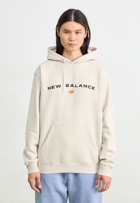 Beige Kapuzenpullover mit Kordelzügen, mit dem "NEW BALANCE"-Logo in Schwarz und Orange über der Brust, Kängurutasche und gerippten Bündchen.