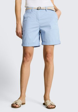 Vrouw draagt lichtblauwe shorts met opgerolde zoom, beige gevlochten riem en witte slides, staand met één hand in haar zak.