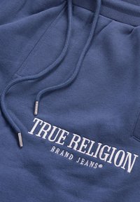 Blue fabric with white embroidered "TRUE RELIGION BRAND JEANS" text and matching blue drawstrings with metal tips.