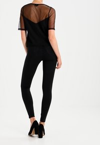 Top nero a maniche corte con schiena in rete trasparente, abbinato a jeans skinny neri e scarpe con tacco alto nere, visto da dietro.