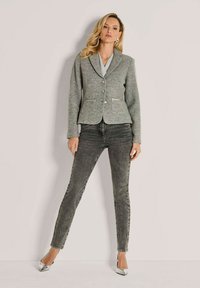 Vrouw staat tegen de muur, gekleed in een grijze blazer, een grijs topje, donkergrijze skinnypants en zilveren puntige hakken.