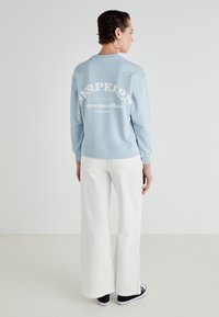 Lichtblauwe hoodie met witte tekst "Skopelos" en "oceankusten." Gecombineerd met witte wijde broek en zwarte sneakers.