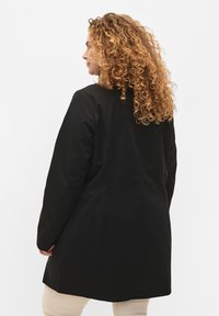 Black long coat with a structured design, featuring a smooth fabric texture and minimalistic silhouette. Back view emphasizes simplicity.