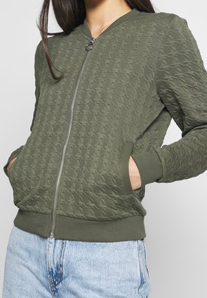 Mujer que lleva una chaqueta acolchada de cremallera de color verde oliva con bolsillos, combinada con vaqueros de mezclilla azul claro.